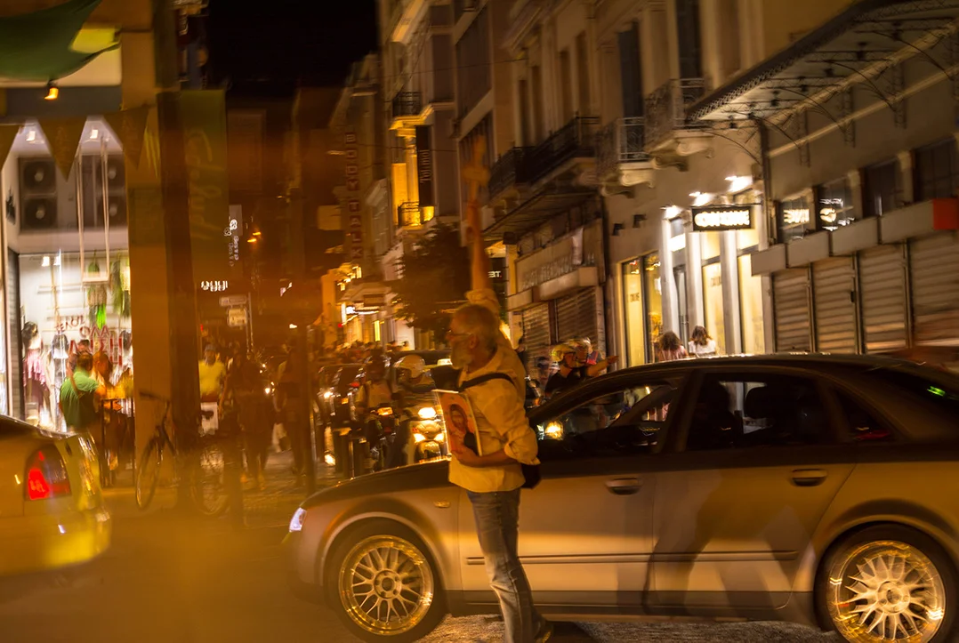 Man holding a cross and an orthodox icon in the middle of a street in Athens surrounded by cars and crowds in the pedestrian street